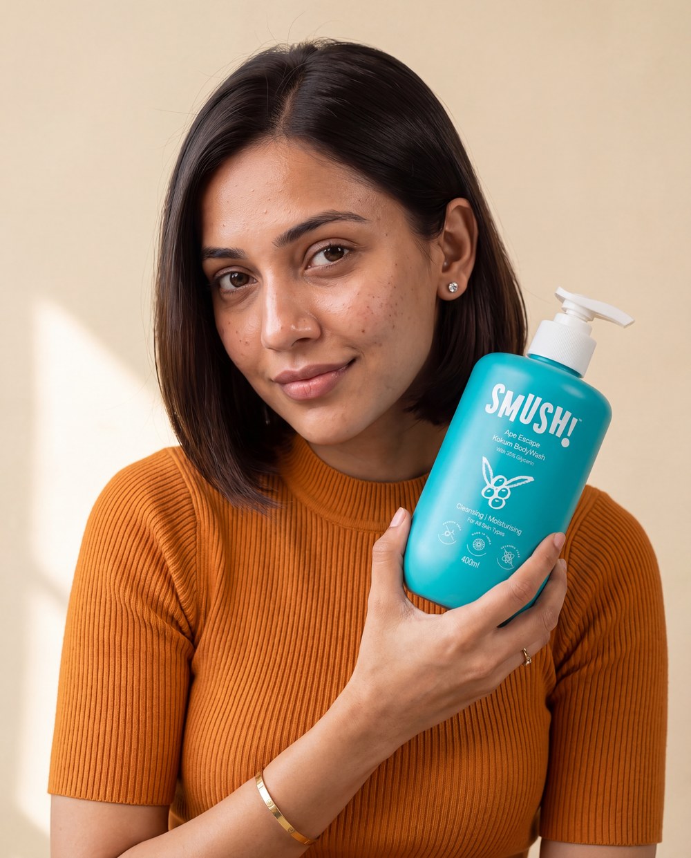 A woman with a sleek bob in a tangerine knit top holds the teal SMUSH! Ape Escape Kokum Body Wash beside her face against a cream wall