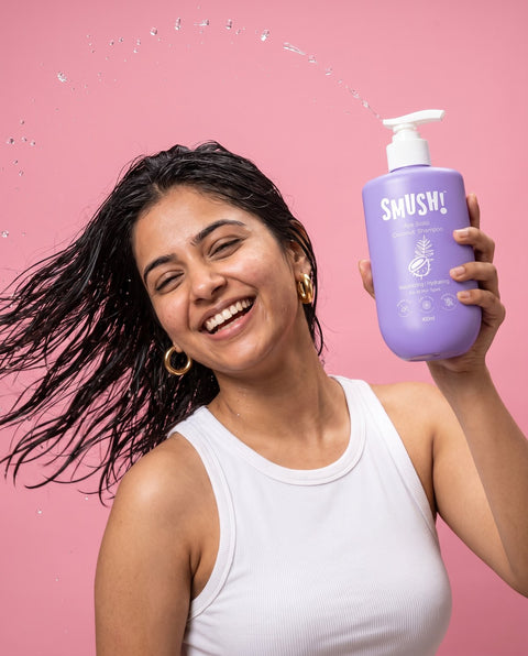 Woman mid hair-flip holding Ape Scalp Coconut Shampoo on a pink backdrop