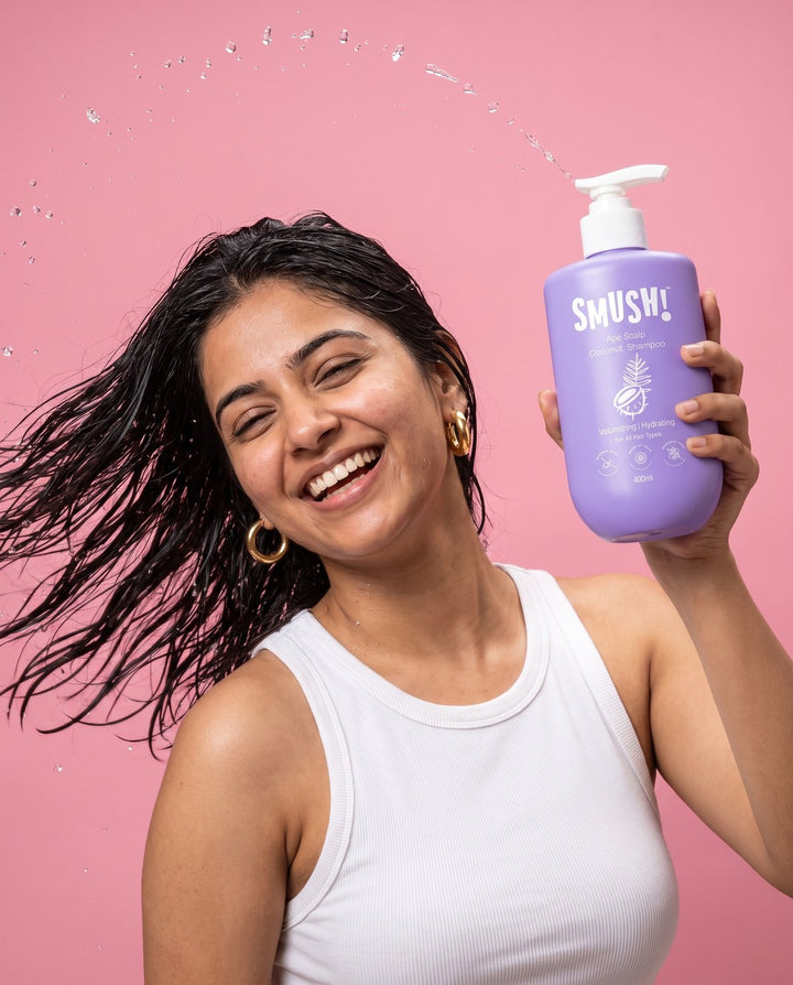 Woman mid hair-flip holding Ape Scalp Coconut Shampoo on a pink backdrop