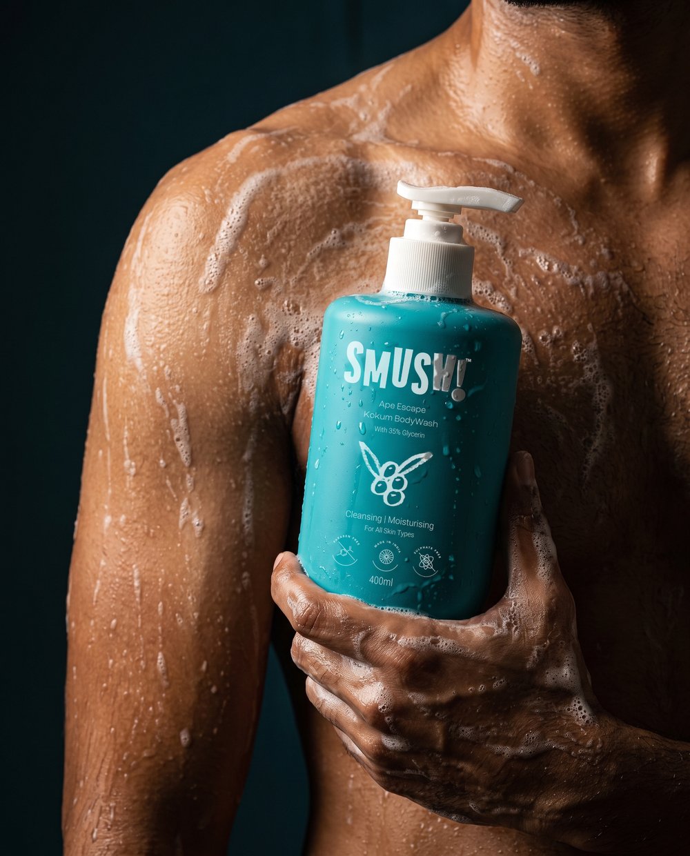 Close-up of a man's wet shoulder and chest streaked with lather, his hand holding the teal SMUSH! Ape Escape Kokum Body Wash, on a dark teal ground