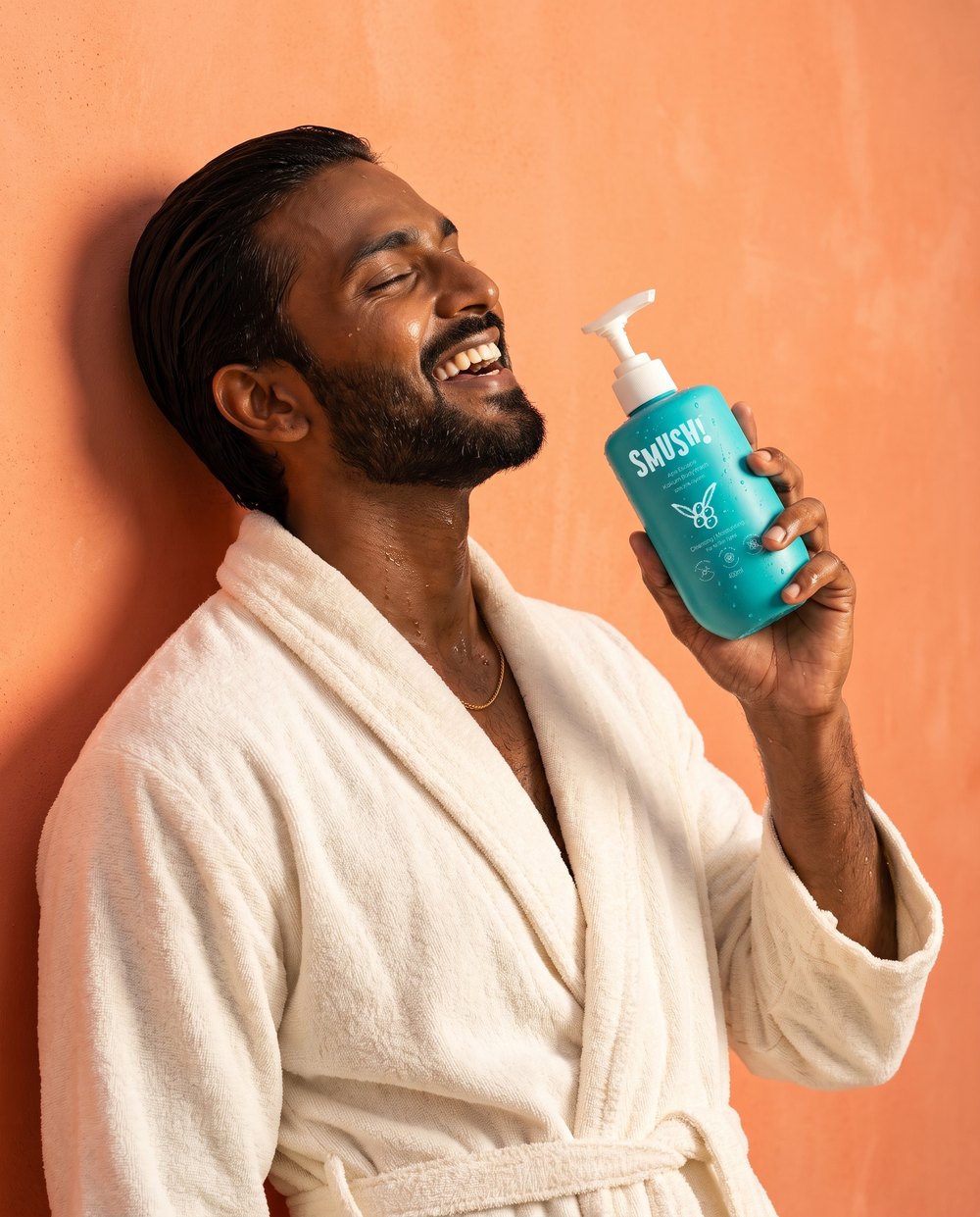 A bearded man in a white bathrobe laughs with his eyes closed and holds the teal SMUSH! Ape Escape Kokum Body Wash against a coral limewash wall