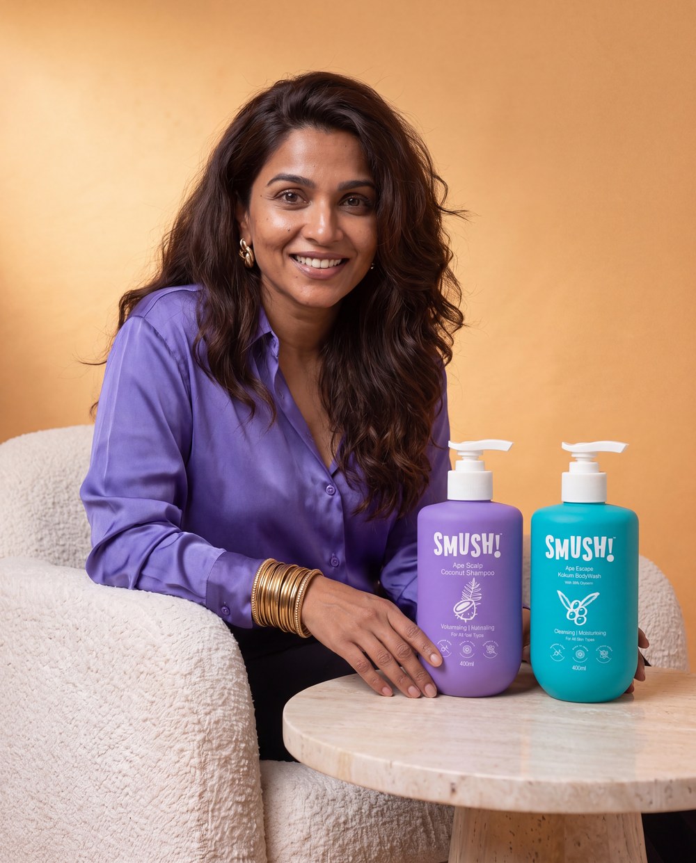 A woman with long wavy hair in a lavender-purple satin shirt smiles beside the purple SMUSH! shampoo and teal SMUSH! body wash standing on a travertine side table, against a saffron wall