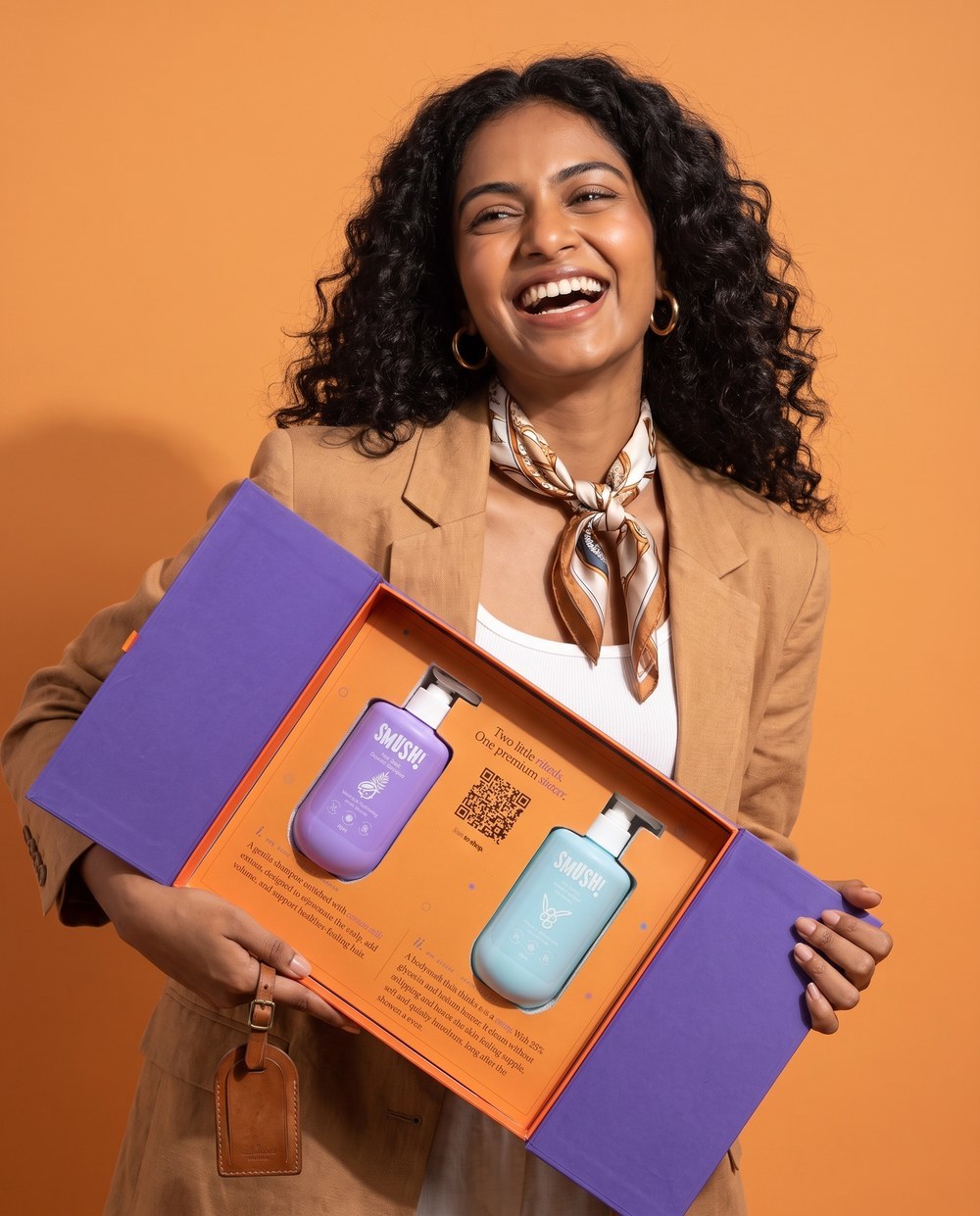 A woman with curly hair in a camel blazer laughs as she holds the open SMUSH! gift box, the shampoo and body wash standing in the orange tray, against an orange wall