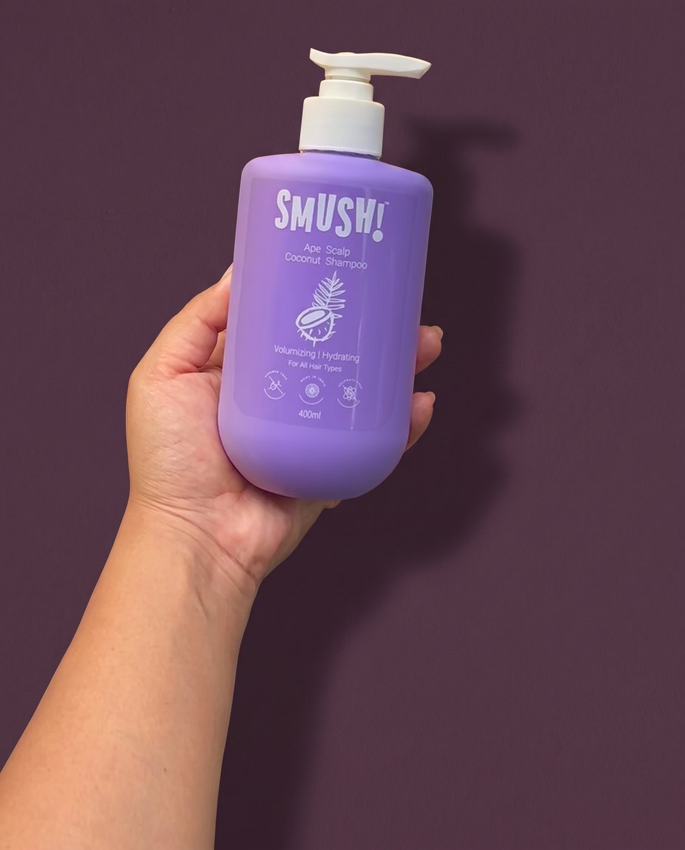 A hand cradles the purple SMUSH! Ape Scalp Coconut Shampoo, the 400 ml bottle as long as the hand, against a deep aubergine wall