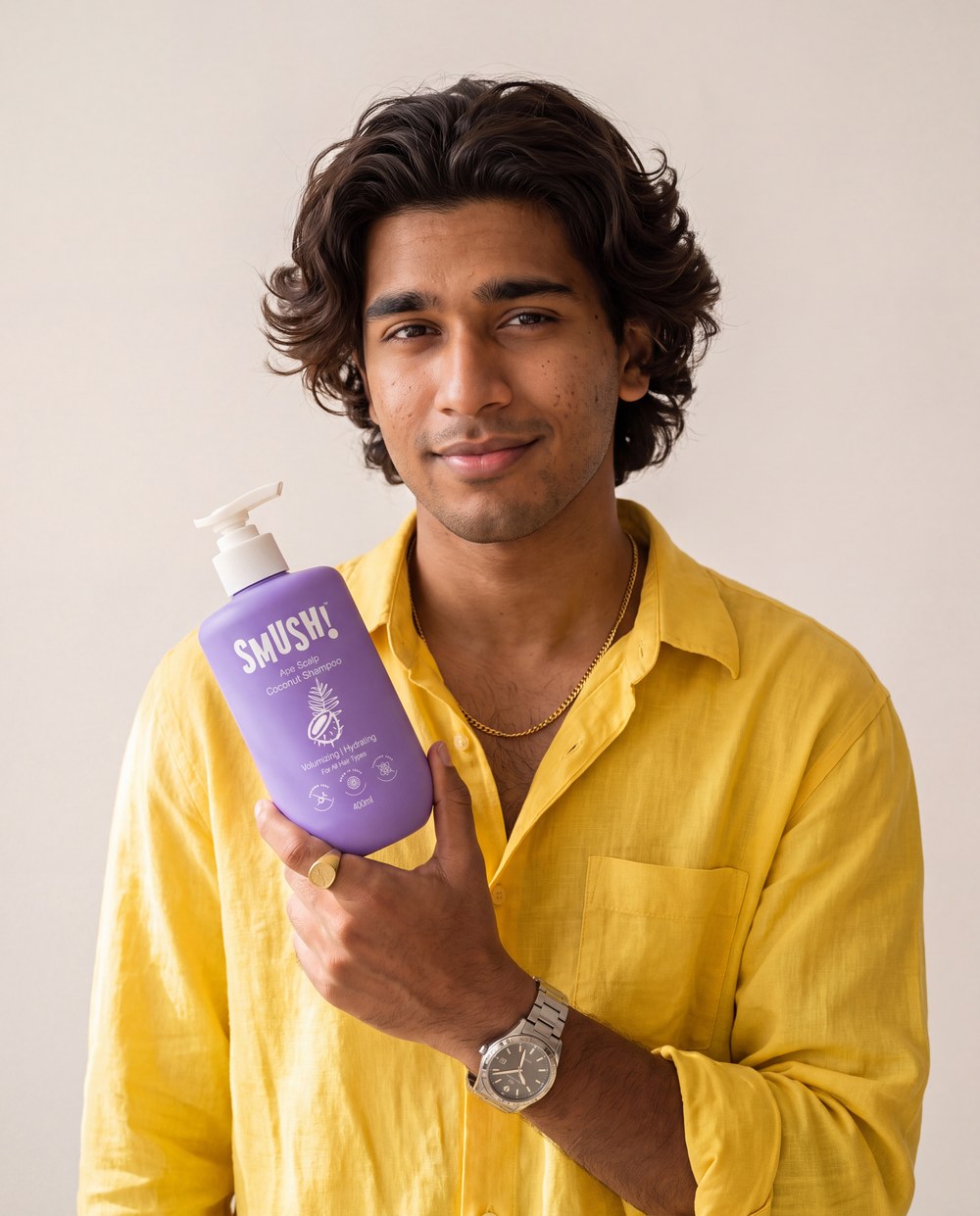 A young man with thick wavy hair in a sunflower-yellow linen shirt holds the purple SMUSH! Ape Scalp Coconut Shampoo at chest height against a warm white wall
