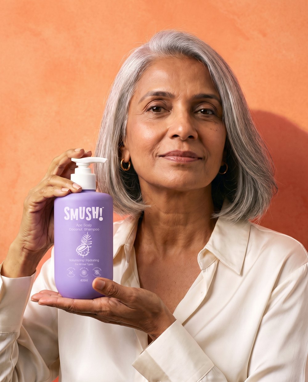 A silver-haired woman in a cream silk shirt smiles and holds the purple SMUSH! Ape Scalp Coconut Shampoo in both hands against a coral limewash wall