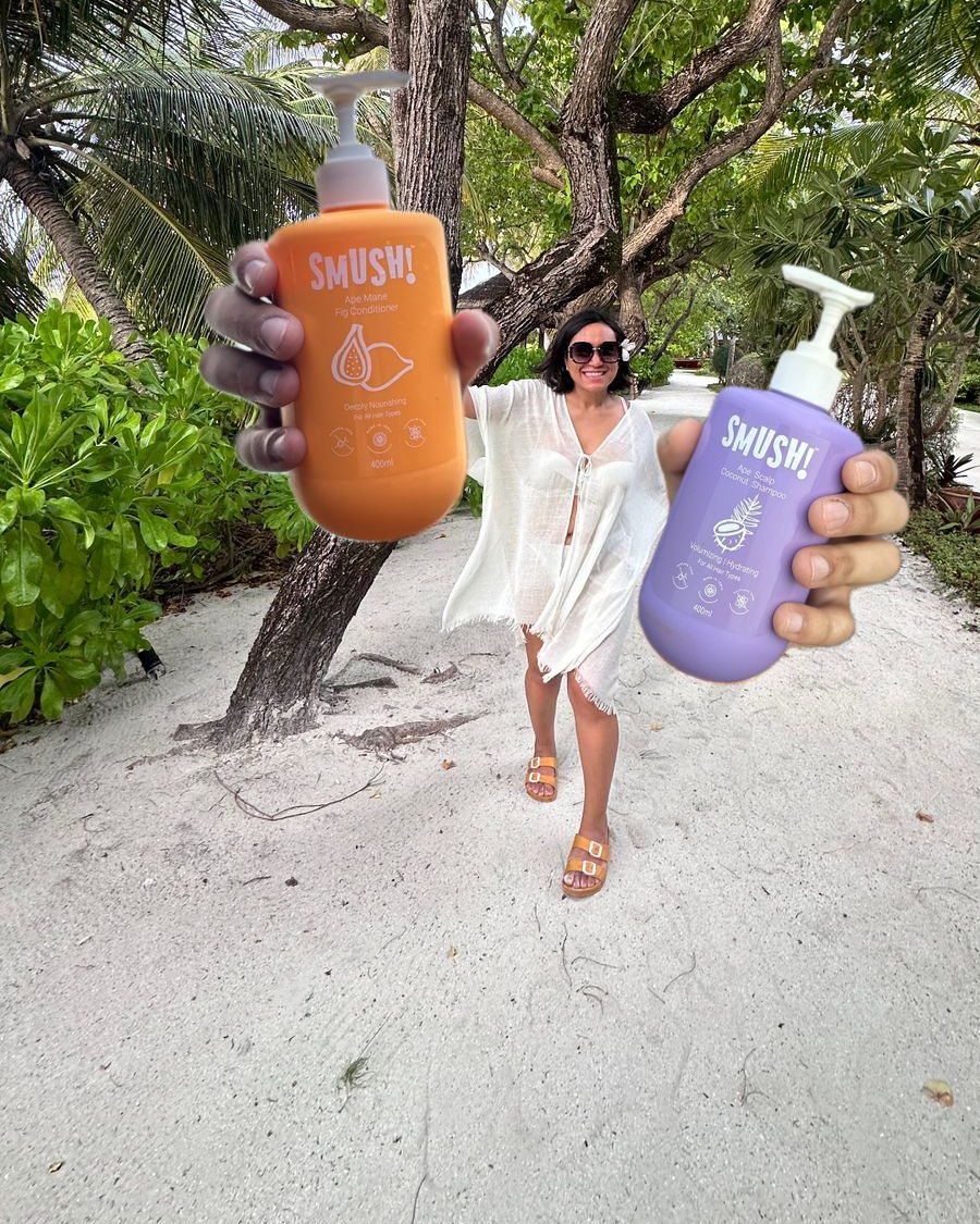 Smriti Mamgain, founder and CEO of SMUSH!, in sunglasses and a white cover-up on a sandy path under trees, holding two SMUSH! bottles out to the camera, the orange conditioner and the purple Ape Scalp Coconut Shampoo