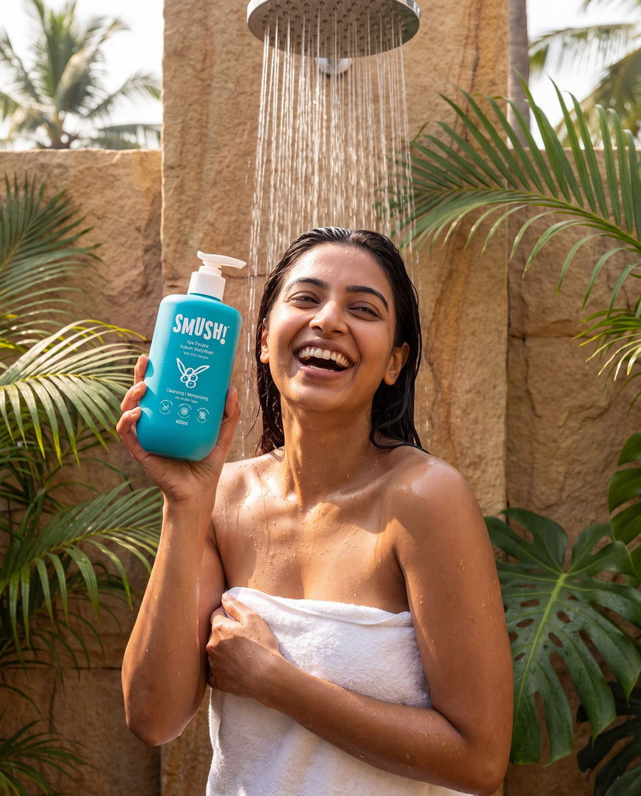 A woman wrapped in a white towel laughs under an outdoor rain shower, holding the teal SMUSH! Ape Escape Kokum Body Wash, palms and a sandstone wall behind