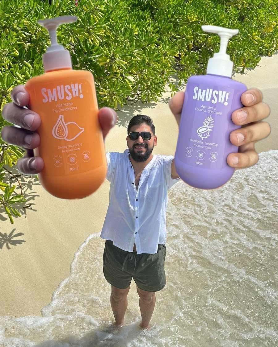 Sahil Khanna, co-founder of SMUSH!, in sunglasses and a white shirt, standing ankle-deep at the water's edge, holding two SMUSH! bottles out to the camera, the orange conditioner and the purple Ape Scalp Coconut Shampoo