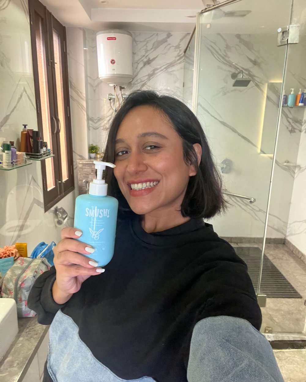 A woman with a short bob smiles in her marble bathroom, holding the teal SMUSH! Ape Escape Kokum Body Wash beside her face
