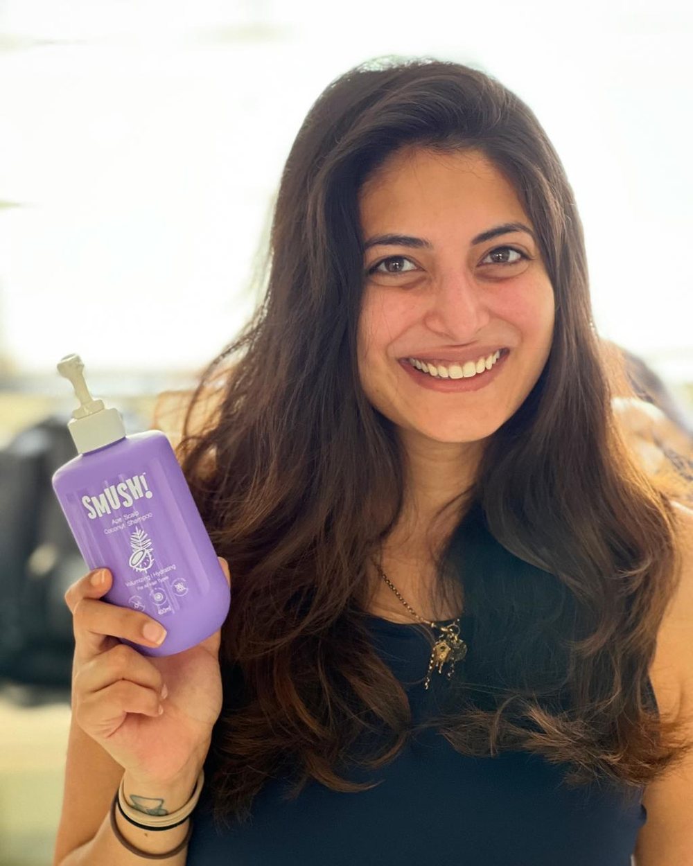 A woman with long wavy hair smiles widely and holds up the purple SMUSH! Ape Scalp Coconut Shampoo