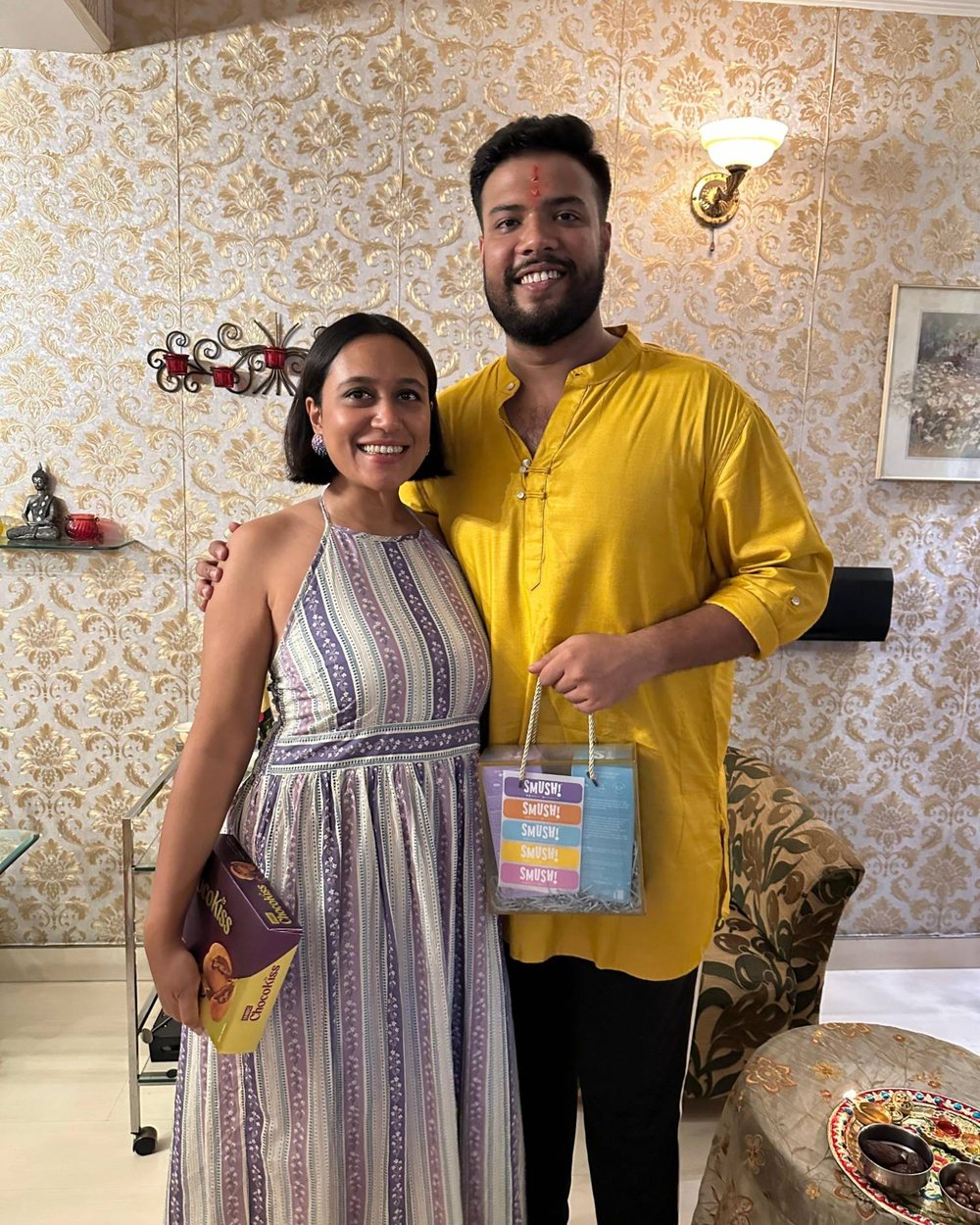 A woman in a striped dress and a man in a yellow kurta pose at home, he holds a SMUSH! gift bag