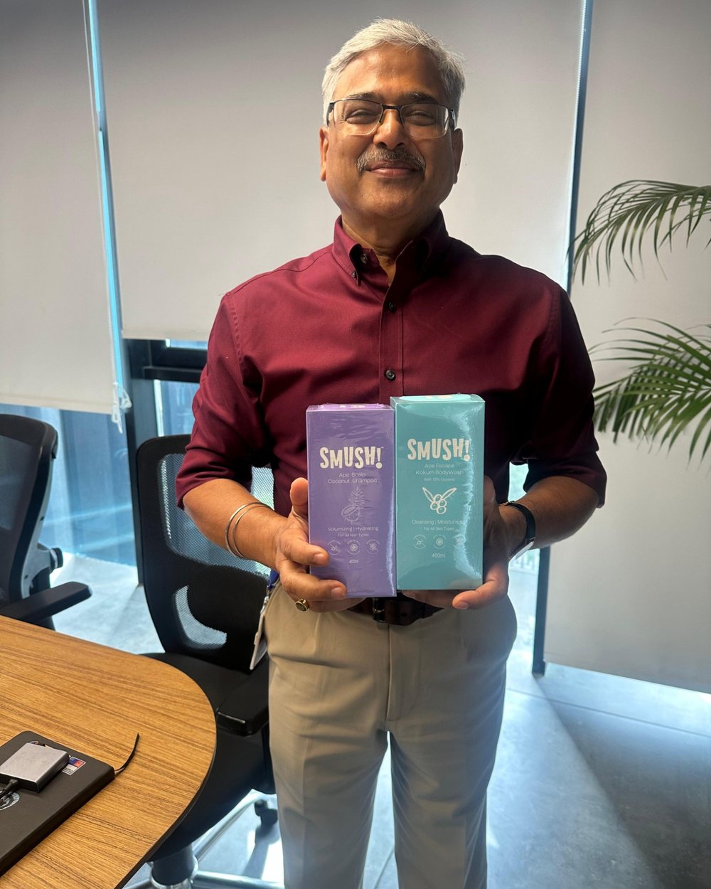 A grey-haired man in a maroon shirt holds the boxed SMUSH! shampoo and body wash side by side in an office