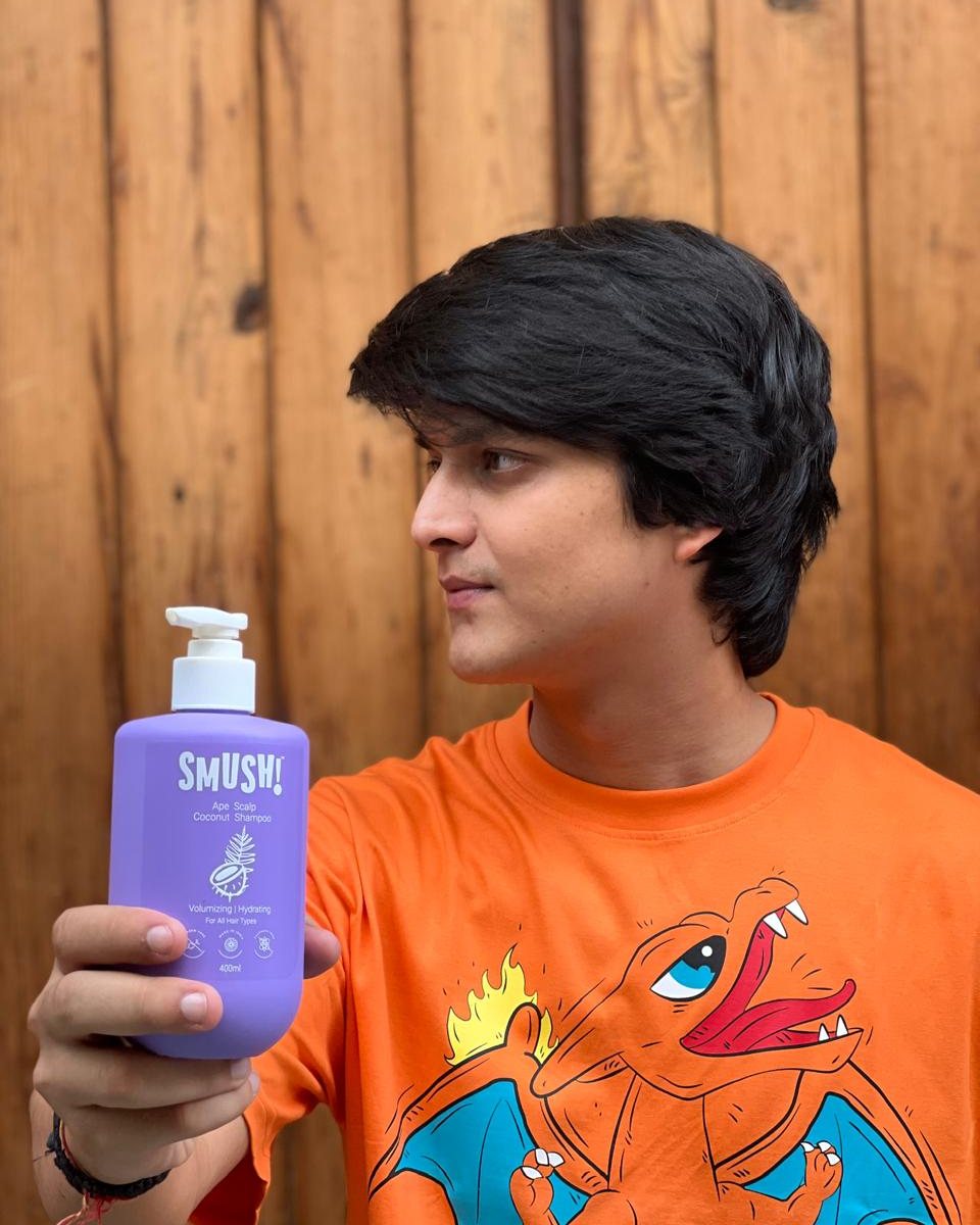 A young man in an orange cartoon T-shirt holds out the purple SMUSH! Ape Scalp Coconut Shampoo against a wooden wall