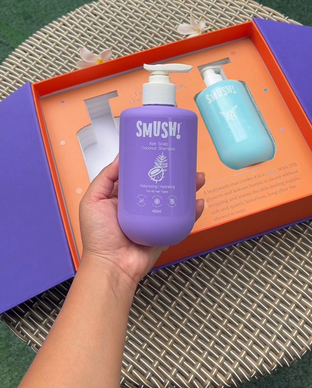 A hand lifts the purple SMUSH! Ape Scalp Coconut Shampoo out of the open gift box, the teal body wash still standing in the orange tray