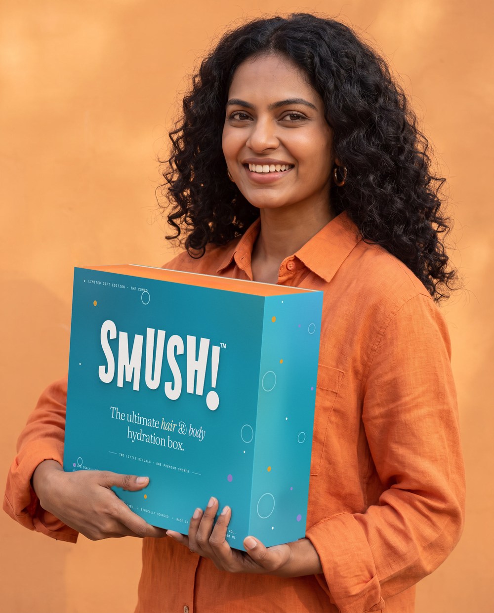 A smiling woman with curly hair in a tangerine linen shirt holds the SMUSH! gift box in its teal printed sleeve, the open top showing the orange box inside