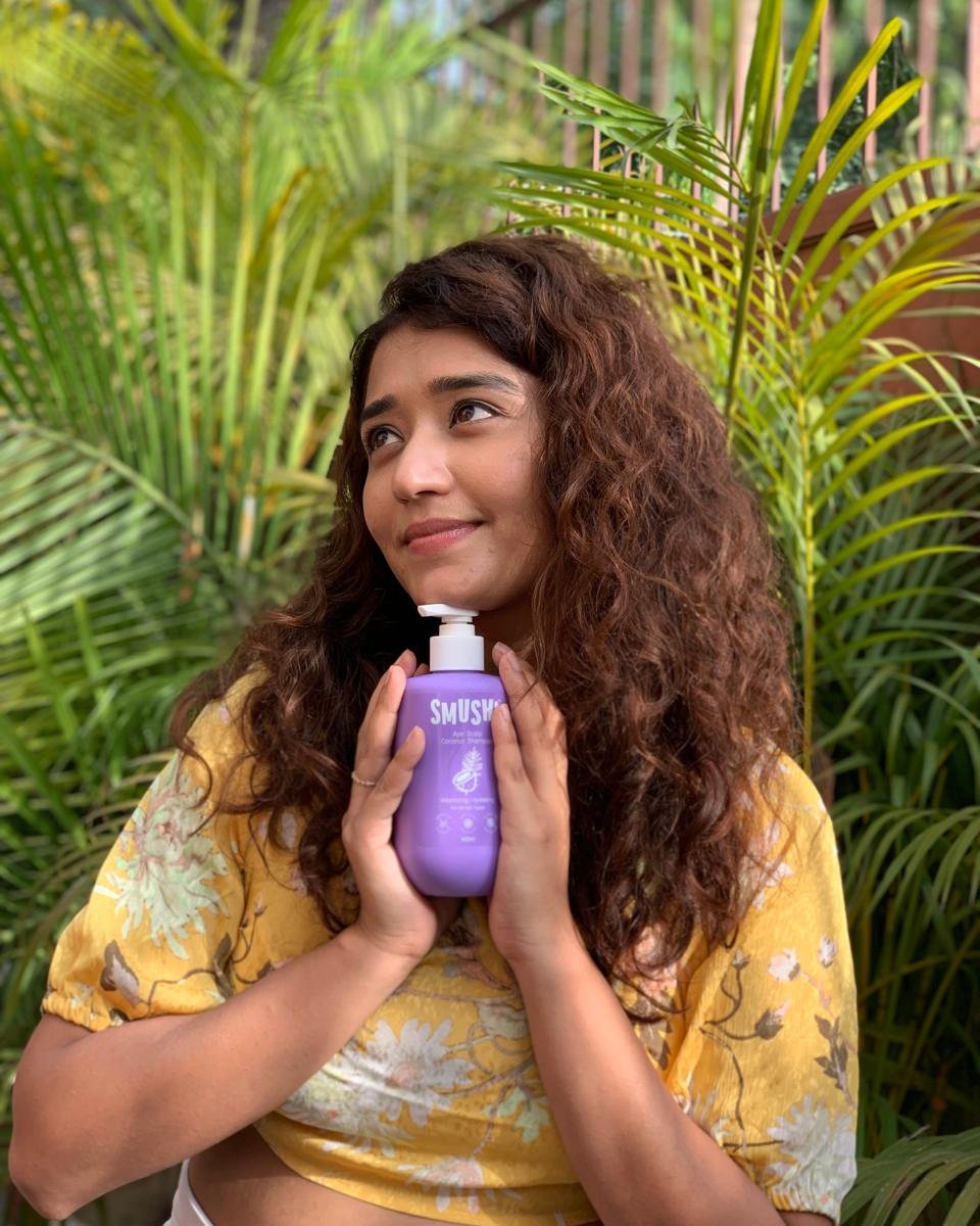 A woman with long curly hair in a yellow floral top hugs the purple SMUSH! shampoo to her chest in front of palm leaves