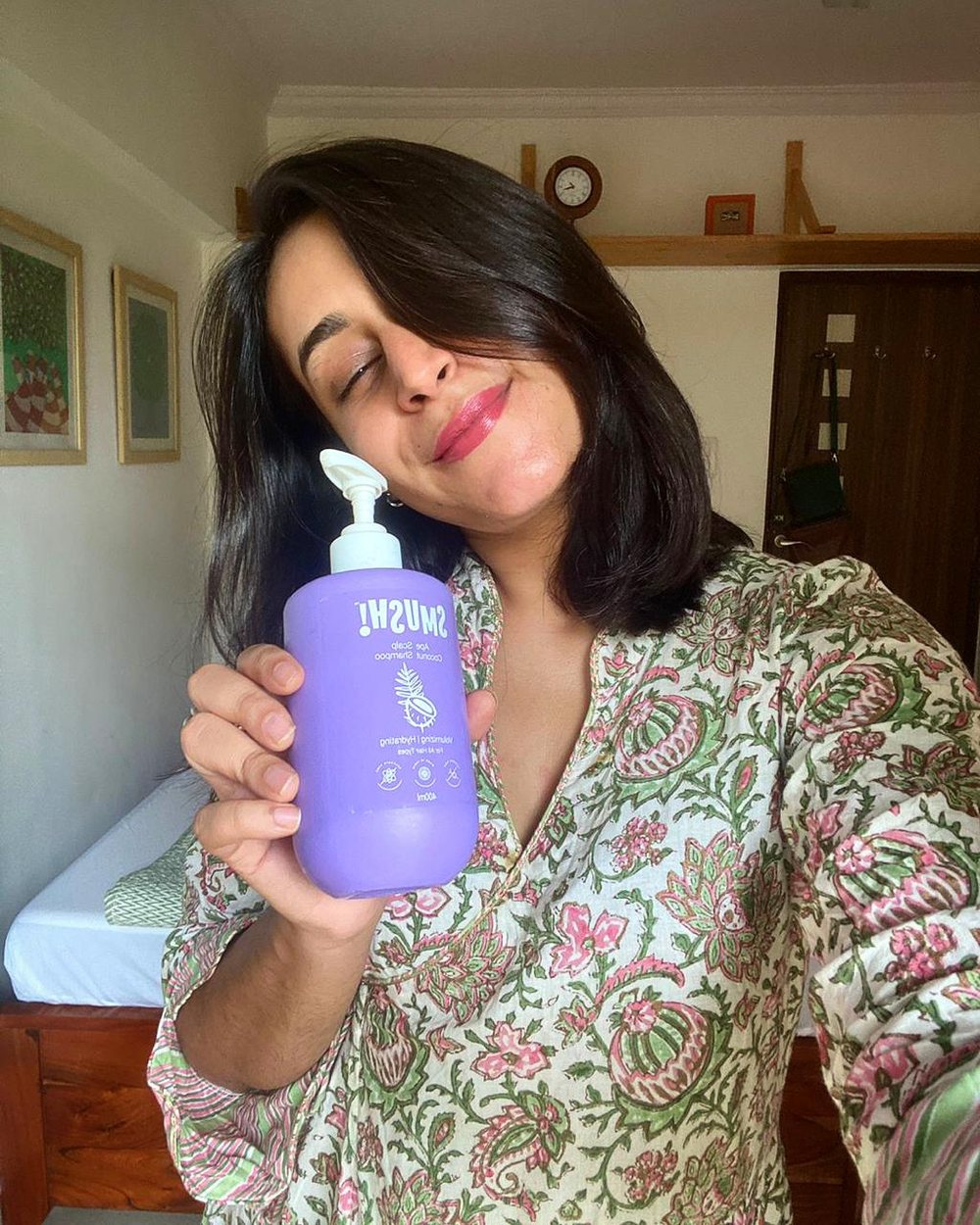 A woman in a floral kurta tilts her head and smiles, holding the purple SMUSH! shampoo beside her face