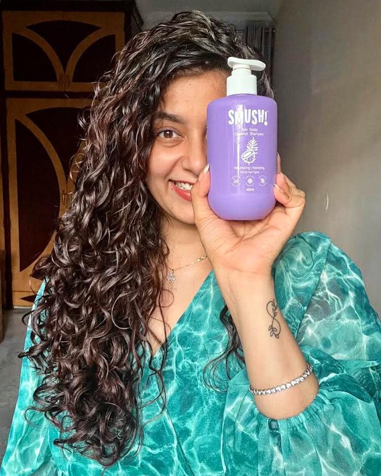 A woman with long curly hair in a sea-green top holds the purple SMUSH! Ape Scalp Coconut Shampoo over one eye and smiles